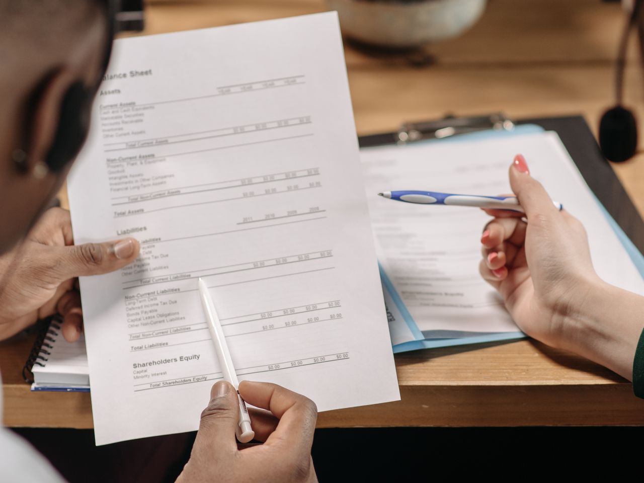 Dos personas revisan un balance financiero y otros documentos sobre una mesa de trabajo.